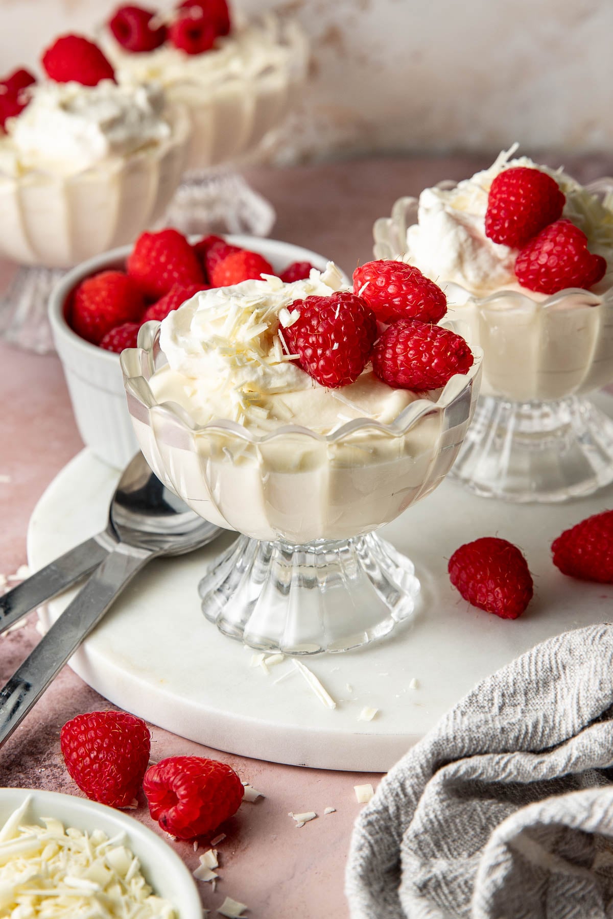 Angled hero shot of white chocolate mousse served in scalloped glass dessert cups topped with whipped cream, raspberries, and white chocolate shavings. Fresh raspberries, dessert spoons, and a linen towel are styled around the desserts on a pink tabletop.