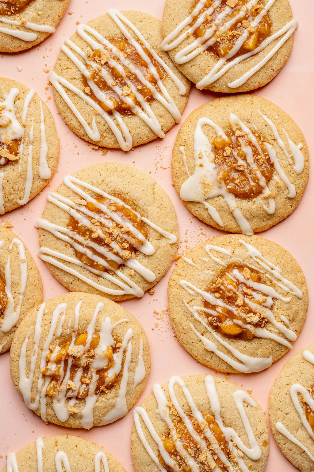 Overhead image of peach cobbler cookies arranged on a blush pink background. Each cookie is topped with peach cobbler filling, white glaze drizzle, and crushed graham cracker crumbs for a cobbler-inspired finish.