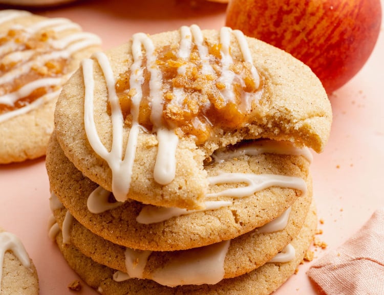 Close-up image of a stack of peach cobbler cookies drizzled with white glaze and topped with peach filling and graham cracker crumbs. The top cookie has a bite taken out, showing the soft and chewy texture inside, with fresh peaches and more cookies blurred in the background.