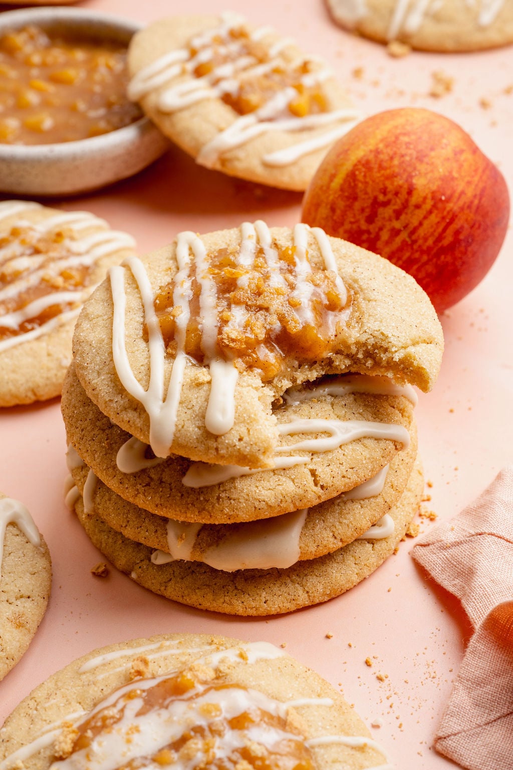 Close-up image of a stack of peach cobbler cookies drizzled with white glaze and topped with peach filling and graham cracker crumbs. The top cookie has a bite taken out, showing the soft and chewy texture inside, with fresh peaches and more cookies blurred in the background.