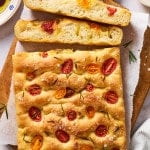Whole tomato rosemary focaccia on a wooden board with sliced pieces showing the light, airy interior, served with olive oil and balsamic vinegar for dipping.