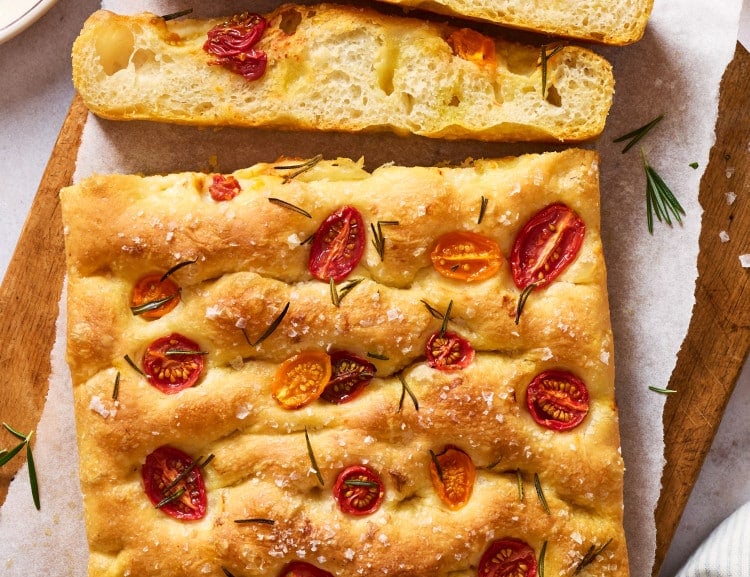 Whole tomato rosemary focaccia on a wooden board with sliced pieces showing the light, airy interior, served with olive oil and balsamic vinegar for dipping.