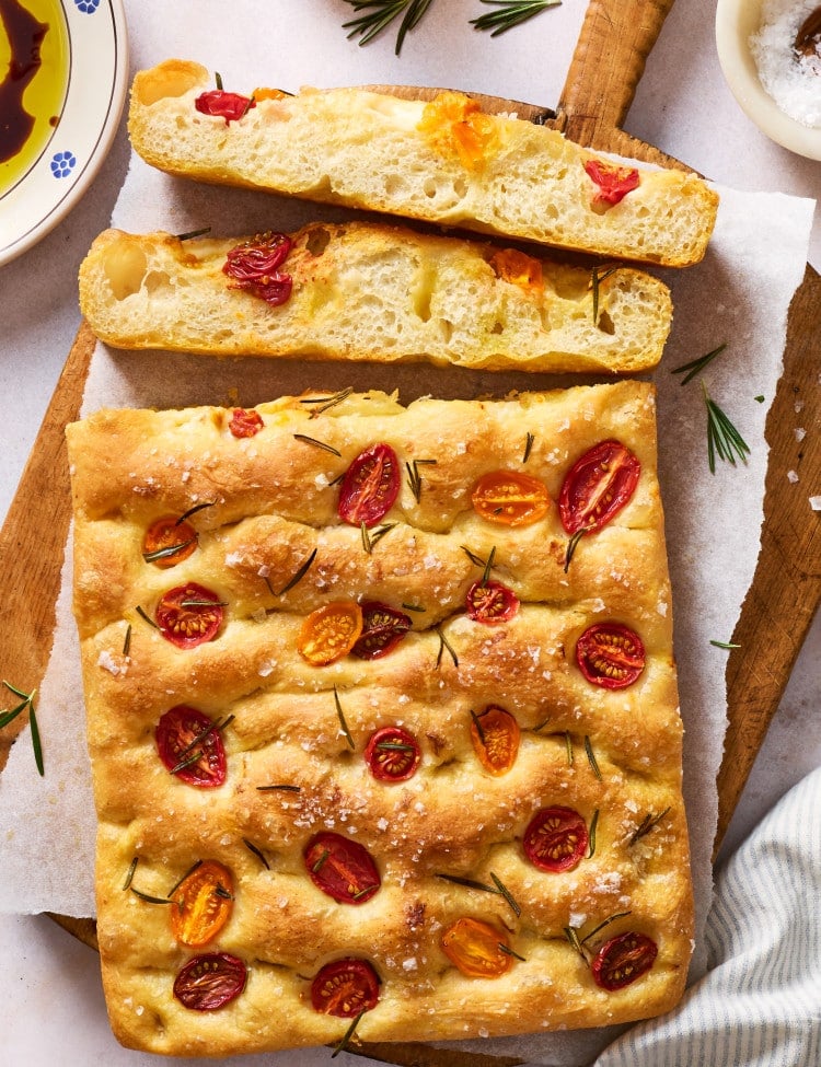 Whole tomato rosemary focaccia on a wooden board with sliced pieces showing the light, airy interior, served with olive oil and balsamic vinegar for dipping.