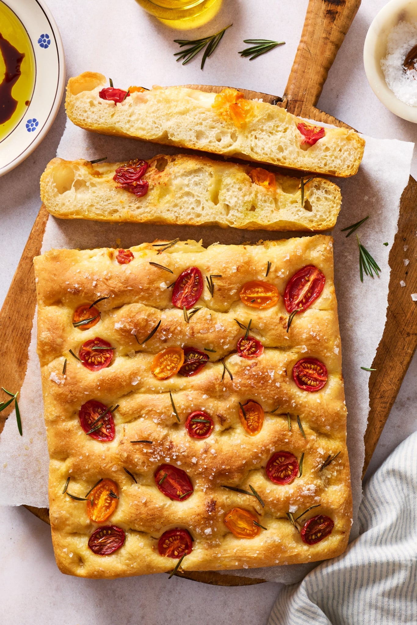 Whole tomato rosemary focaccia on a wooden board with sliced pieces showing the light, airy interior, served with olive oil and balsamic vinegar for dipping.