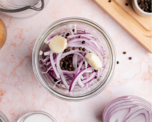 Adding peppercorns, garlic and sliced red onions to large glass jar.