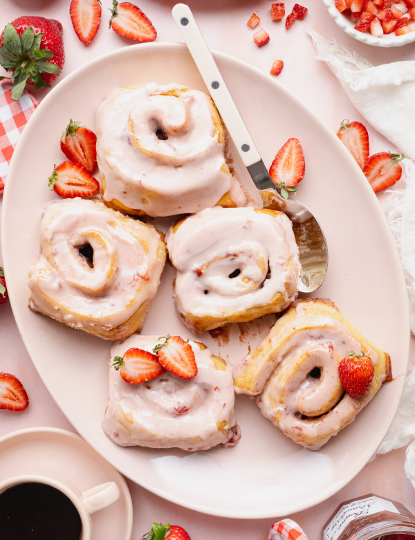 Five strawberry cinnamon rolls on a large oval plate with spoon and extra fresh strawberries.
