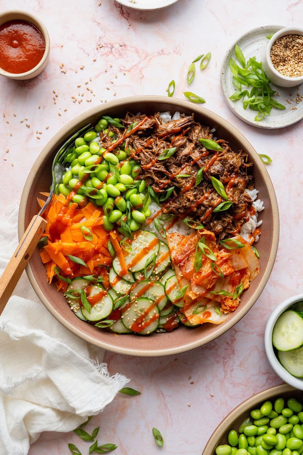 Overhead photo of a Korean-inspired beef bowl topped with tender shredded beef, edamame, cucumber salad, kimchi, shredded carrots, green onions, sesame seeds, and a drizzle of sriracha.