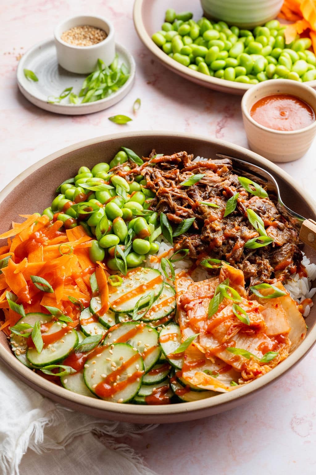 Angled view of a Korean-inspired beef bowl with slow cooker shredded beef served over rice with edamame, cucumber salad, kimchi, carrots, green onions, and sriracha.